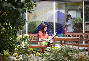 Eine Patientin sitzt auf einer Bank im Park der Rehaklinik Am Kurpark und liest Zeitung.