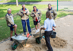 Ein Therapeut mit einer mit Sand gefüllten Schubkarre unterhält sich mit vier Männern, zwei davon halten eine Schaufel.