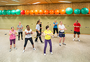 Eine Gruppe von Frauen und Männern macht unter Anleitung eines Therapeuten Gymnastikübungen in einer Turnhalle.